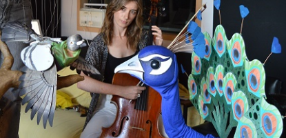 Woman holds cello behind pigeon and peacock puppets
